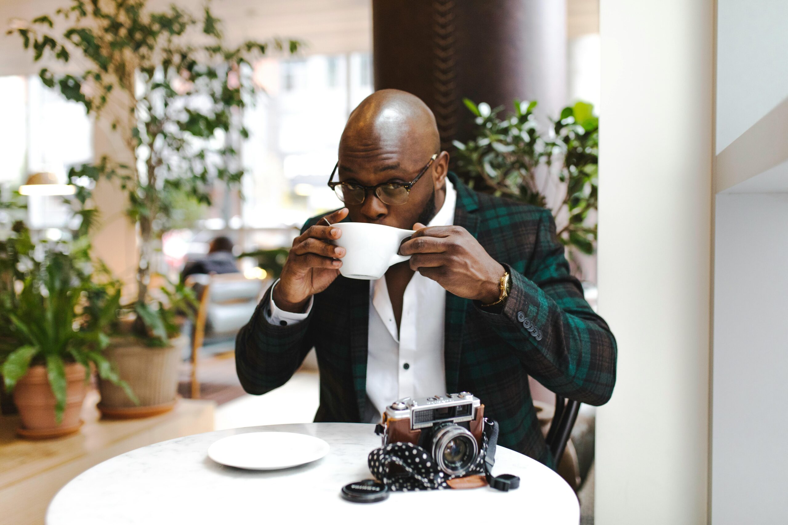 A man sipping coffee in a cafe with a vintage camera on the table. Relaxed indoor setting.